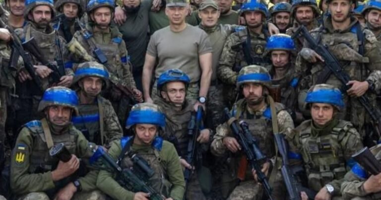 Військовослужбовець у камуфляжі стоїть на тлі руїн, тримаючи зброю, нагадуючи про скандал із наказами відкривати вогонь.